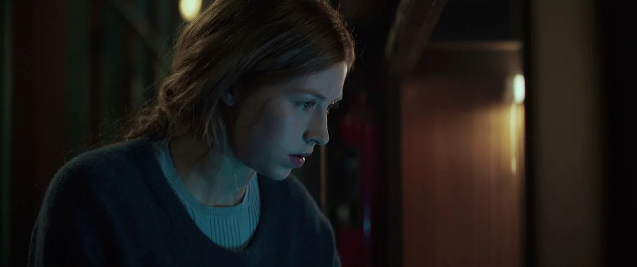 Sea Fever (2019) movie still: closeup — A dimly lit room with shadows obscuring the background details behind the subject. A young woman looks…