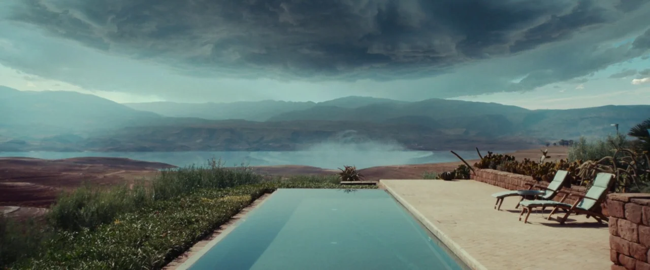 Animalia (2023) movie still: establishing — An infinity pool overlooks a vast arid landscape with a distant lake and mountains. A dark, heavy storm…