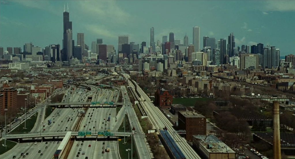 Source Code (2011) movie still: establishing — An elevated wide view shows a sprawling highway interchange near a dense urban core. The skyline…