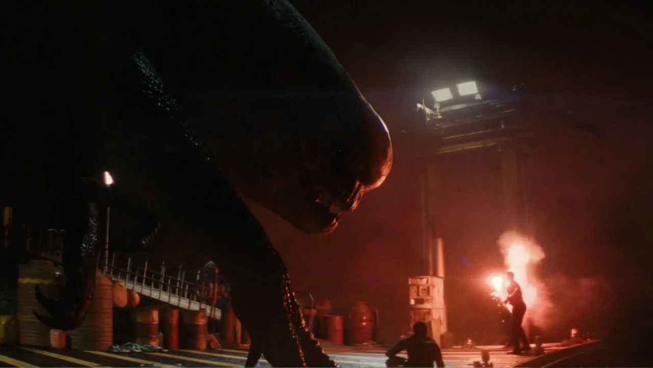 Jurassic World Rebirth (2025) movie still: wide — An industrial pier at night is bathed in the red light of a flare. A large, dark reptilian…