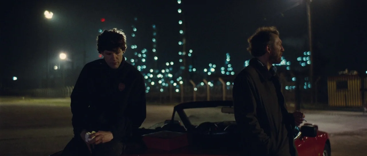 Our Day Will Come (2010) movie still: medium — An industrial lot at night features a glowing refinery plant in the distance. Two men stand beside a…