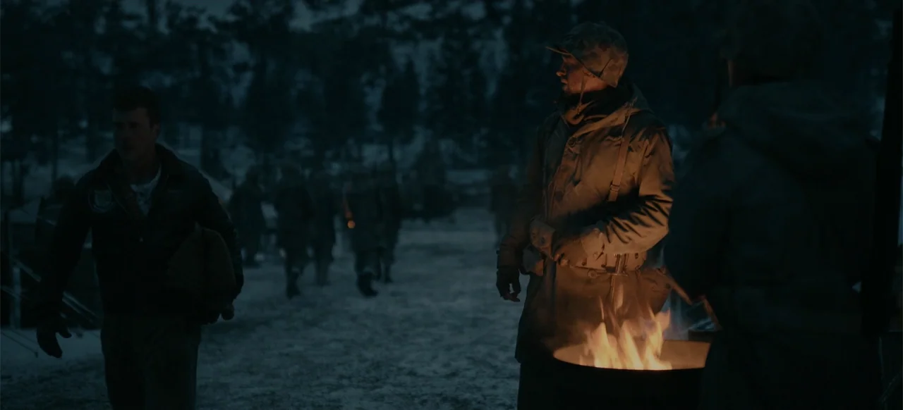 Devotion (2022) movie still: medium — A dark military camp at night with pine trees in the background under moonlight. Two soldiers stand around a…
