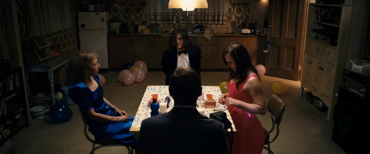The Loved Ones (2009) movie still: medium — A dimly lit kitchen with vintage patterned wallpaper features a table set for dinner. Four people are…