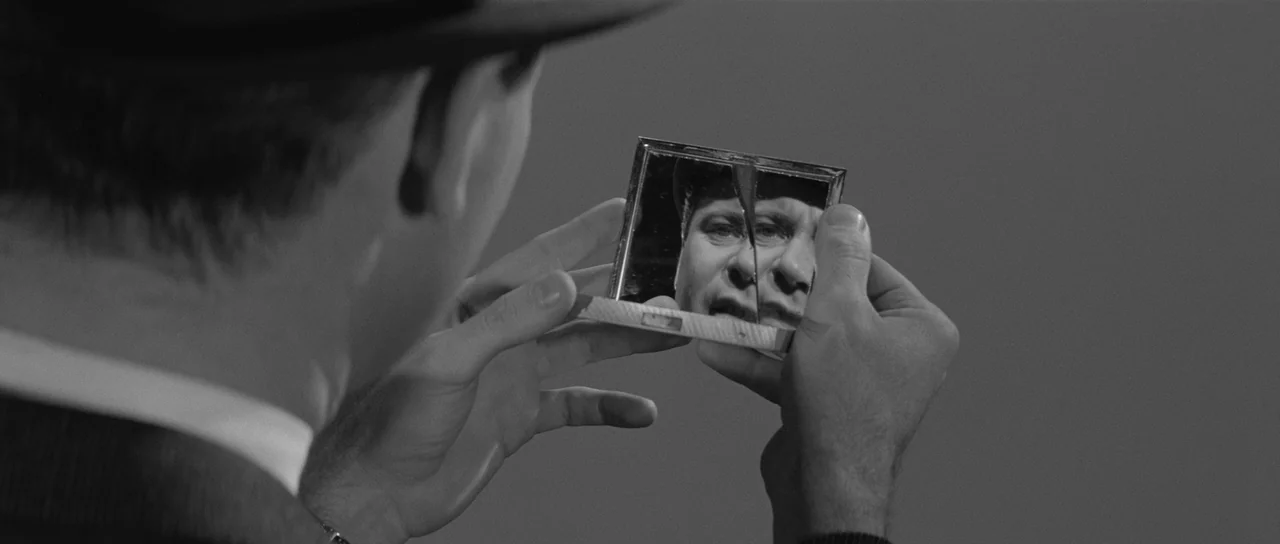 The Apartment (1960) movie still: closeup — A man holds a fractured pocket mirror in his hands to view his face. The background is a solid dark…