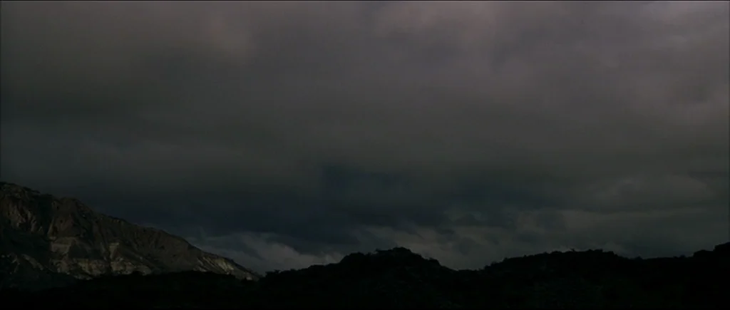 Gerry (2002) movie still: establishing — A dark mountainous landscape sits under a vast, heavy, and overcast grey sky. The scene shows the rugged…