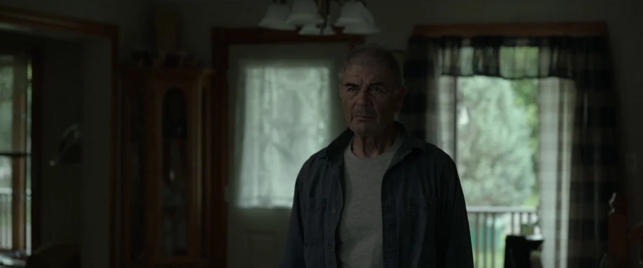 Small Crimes (2017) movie still: medium — A dimly lit room with wood trim and a window with patterned curtains. An older man stands centered in the…
