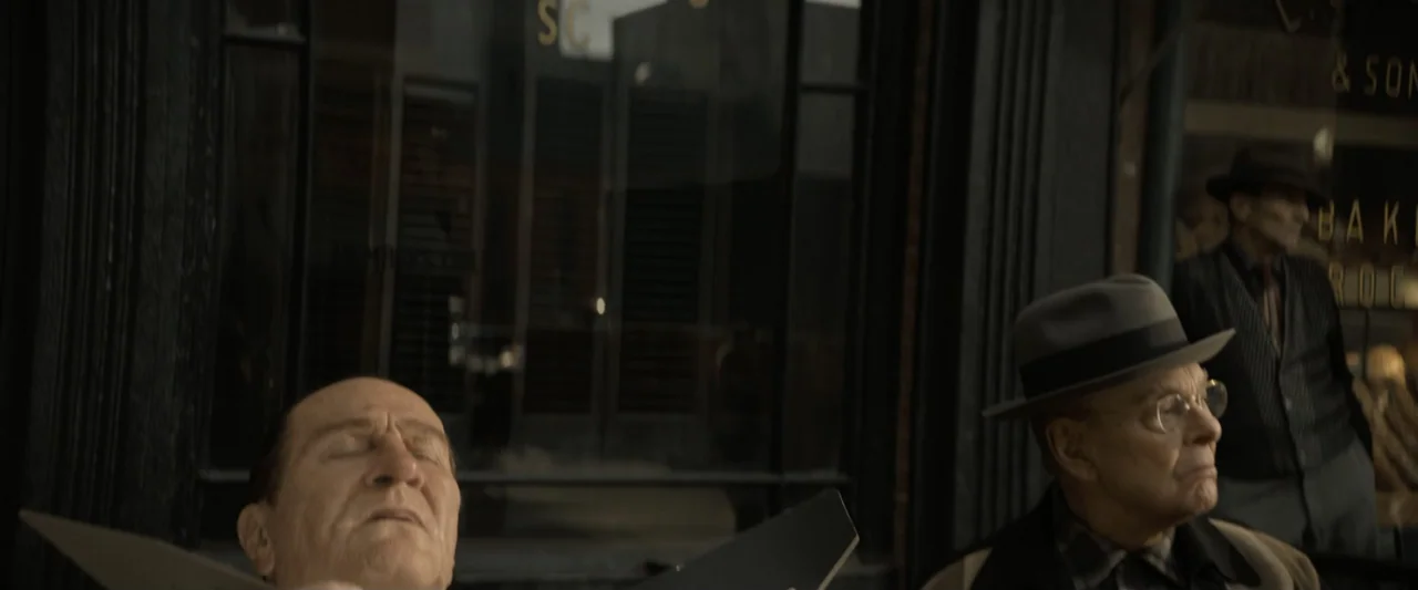 The Alto Knights (2025) movie still: medium — A 1940s urban storefront features dark display windows with gold lettering. Two older men sit together…