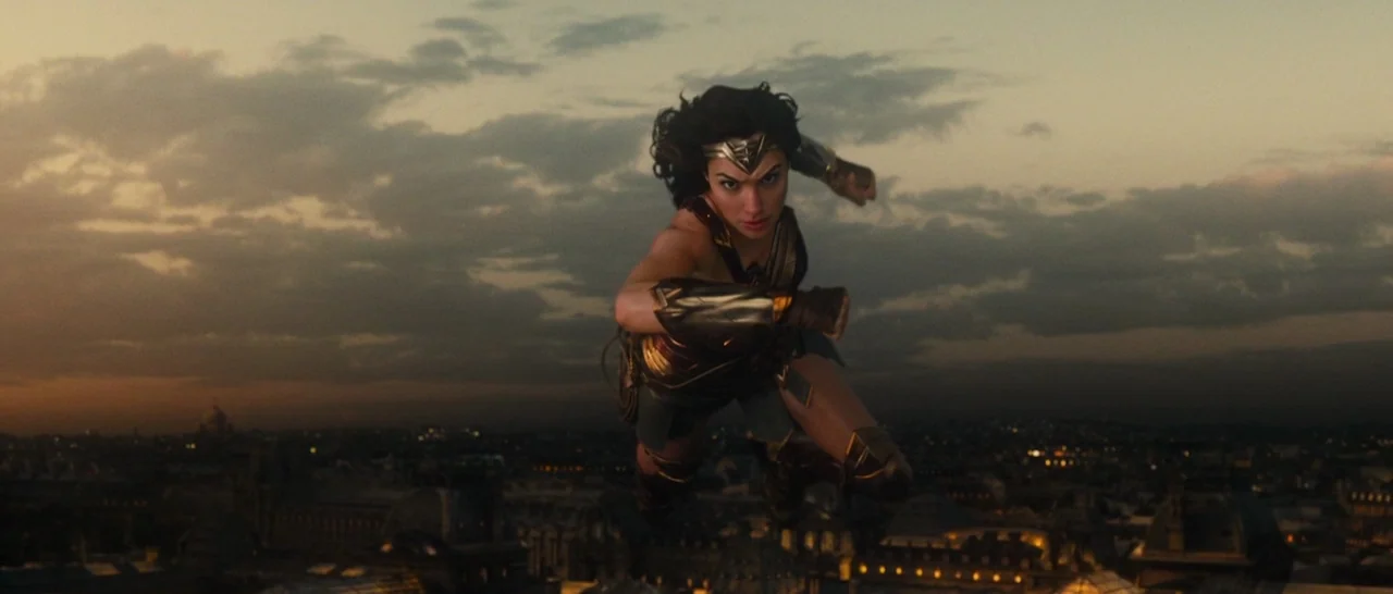 Wonder Woman (2017) movie still: fullbody — A dark city landscape is visible far below against a golden, overcast sunset sky. A female warrior flies…