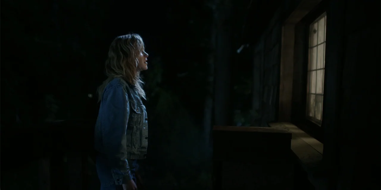 Totally Killer (2023) movie still: medium — A dark outdoor wooden deck at night featuring a single illuminated window in the wall. A young woman in…