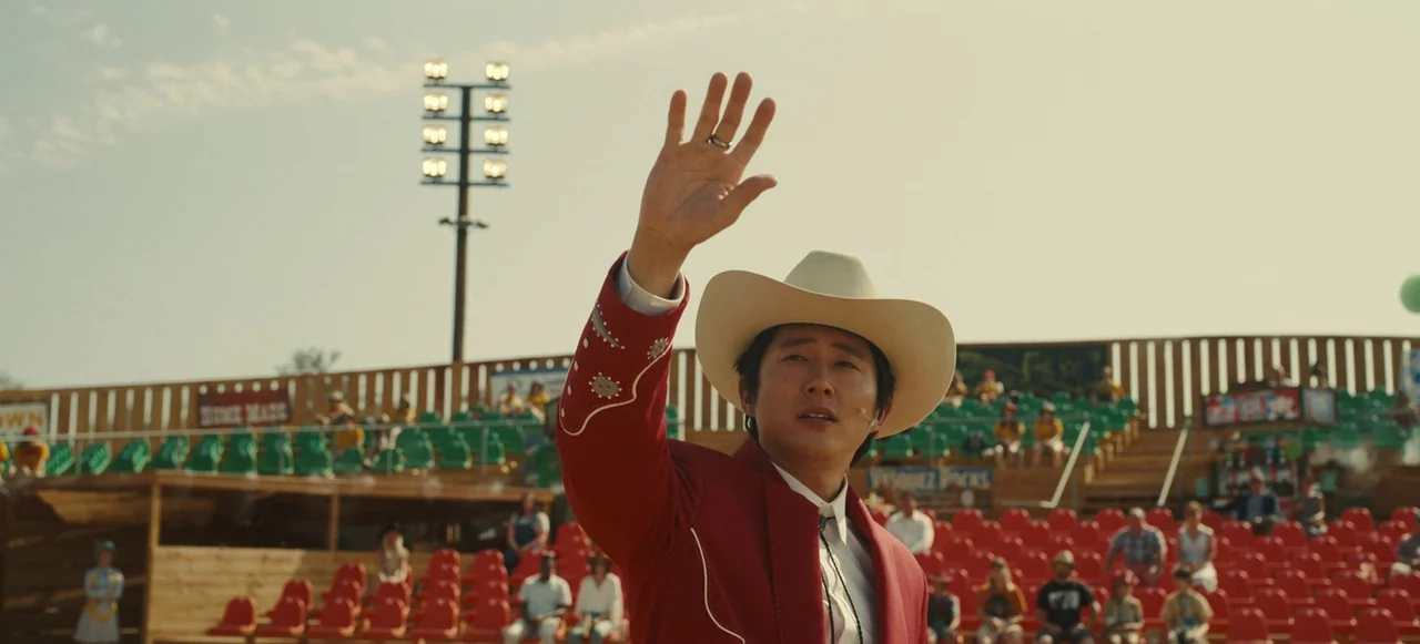 Nope (2022) movie still: medium — An outdoor arena with tiered red and green seating under a bright clear sky. A man in a red western blazer stands…