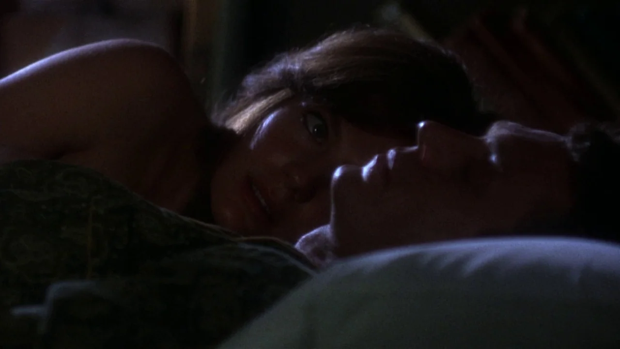 Bullitt (1968) movie still: closeup — A dark bedroom with bedding visible in the foreground. A woman stares at a sleeping man in the dim light.