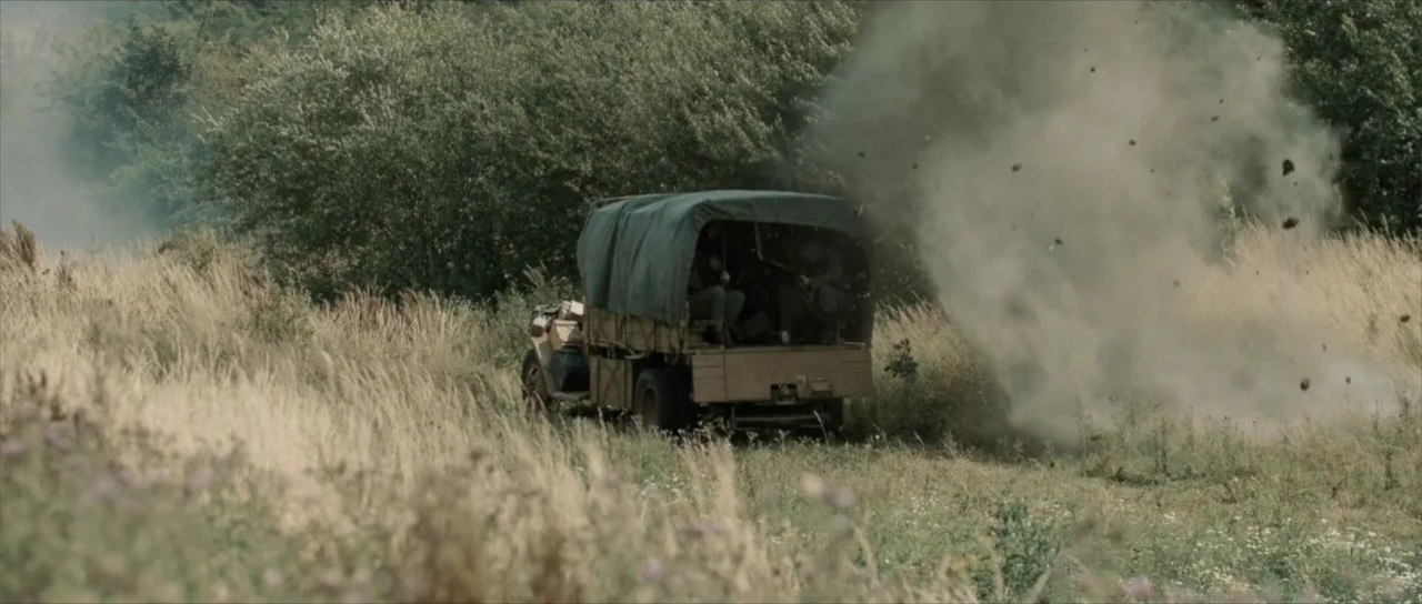 Sin retorno (2009) movie still: wide — An open grassy field contains a military truck with a canvas roof cover. Dust and dirt erupt into the air as…