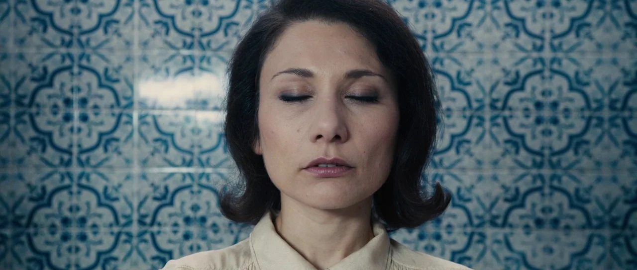 The Duke of Burgundy (2014) movie still: closeup — A wall covered in blue and white ornate patterned tiles forms the background. A woman stands in…