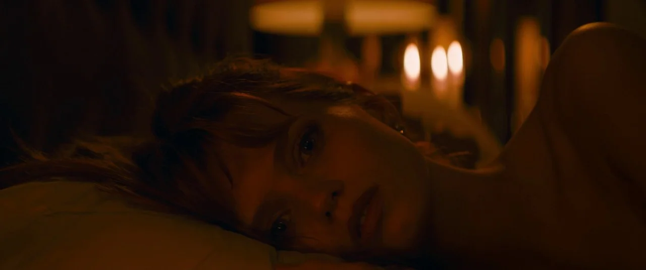Elizabeth Harvest movie still: closeup — A dimly lit space features a woman lying on a white pillow. Blurred artificial lights glow softly in the…