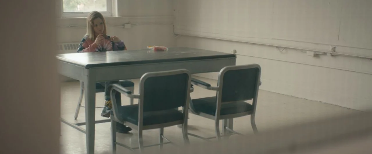 Becky (2020) movie still: wide — A stark room features a gray table and two metal-framed chairs. A girl sits at the table holding an object while…