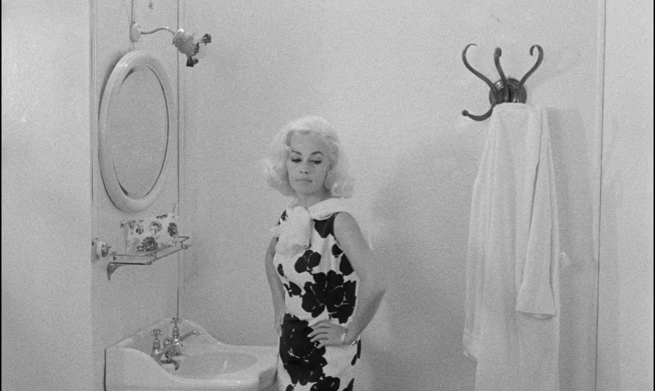 Bay of Angels (1963) movie still: medium — A 1960s bathroom features a simple porcelain sink, a wall mirror, and a bathrobe. A blonde woman stands…