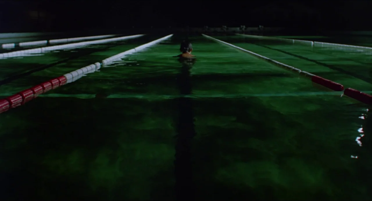 Typhoon Club (1985) movie still: wide — A dark indoor pool features a single person swimming in a lane at night. The water reflects the dim green…