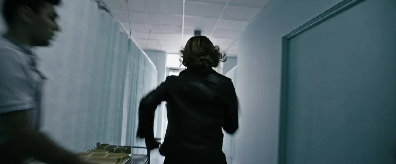 Jolt (2021) movie still: medium — A sterile medical hallway features light-colored walls and hanging privacy curtains on one side. A woman runs past…
