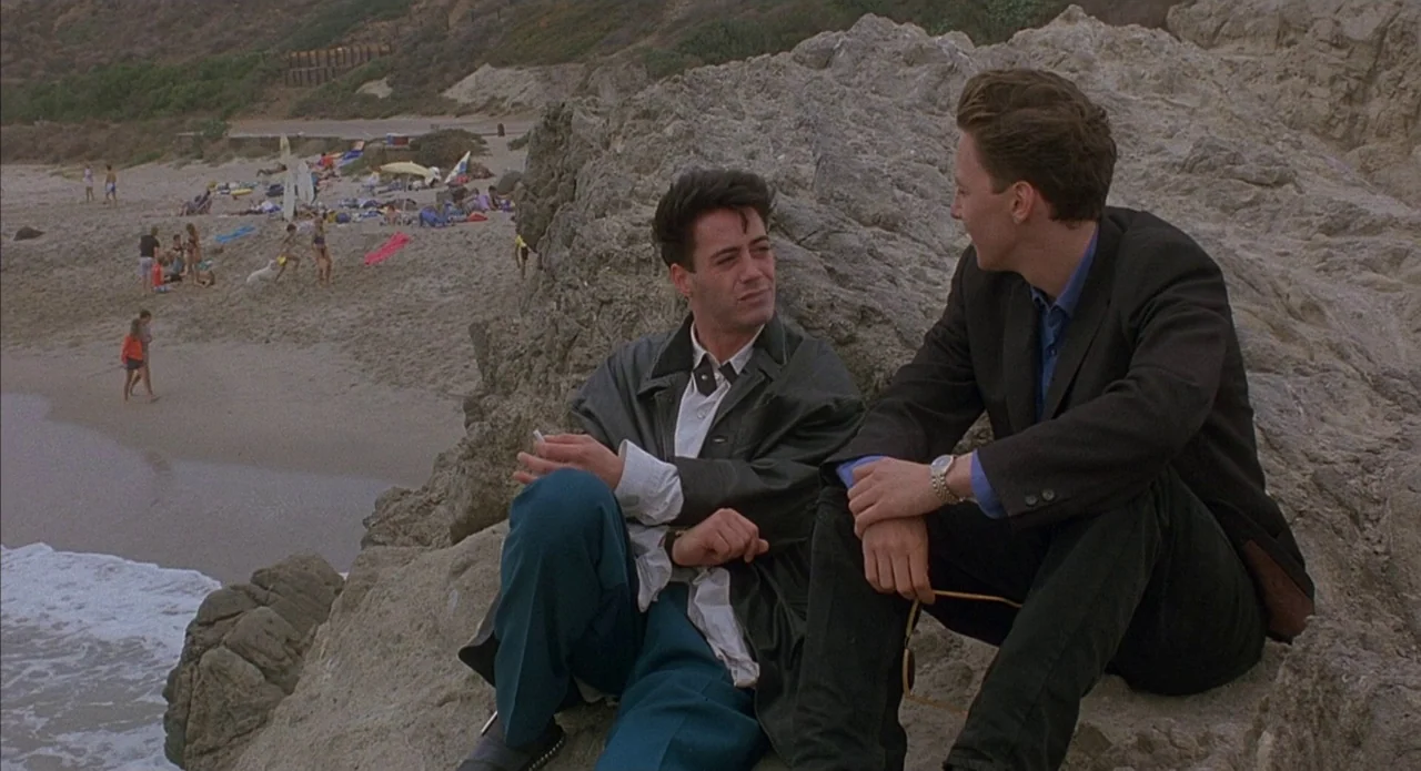 Less Than Zero (1987) movie still: fullbody — Two men sit on a large rocky formation overlooking a populated sandy coastline. They are engaged in…