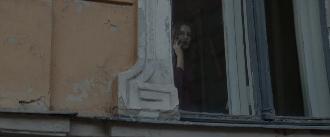La herencia (2010) movie still: medium — A weathered, beige stucco wall with a window reveals a person inside. A woman stands in the dark interior…