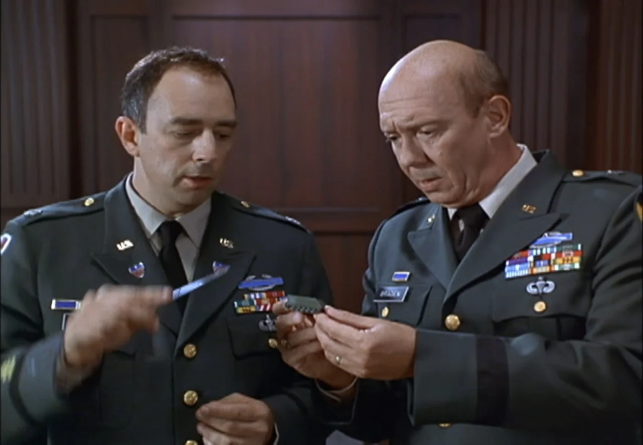 The Pentagon Wars (1998) movie still: medium — Two men in army dress uniforms stand in an office with dark wood paneling. The older man examines a…
