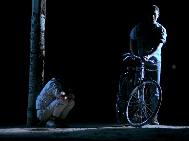 Black Snow (1990) movie still: fullbody — An outdoor area at night features a lone tree and a bicycle illuminated by light. One person sits crouched…