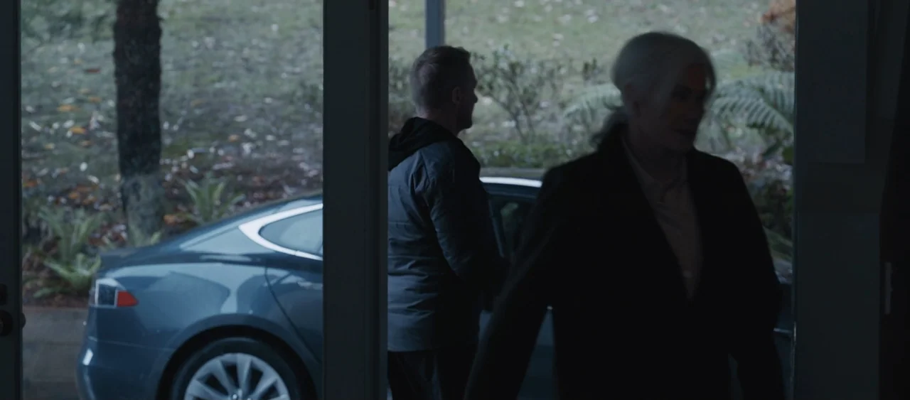 Force of Nature: The Dry 2 (2024) movie still: medium — An outdoor area with a grey sedan parked on a paved surface near greenery. A woman and a man…