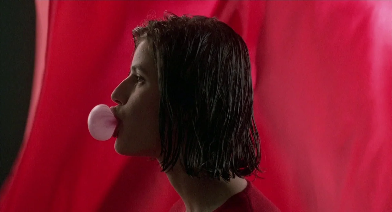 Three Colours: Red (1994) movie still: closeup — A woman stands in profile against a bright, textured red fabric backdrop. She blows a small, round…