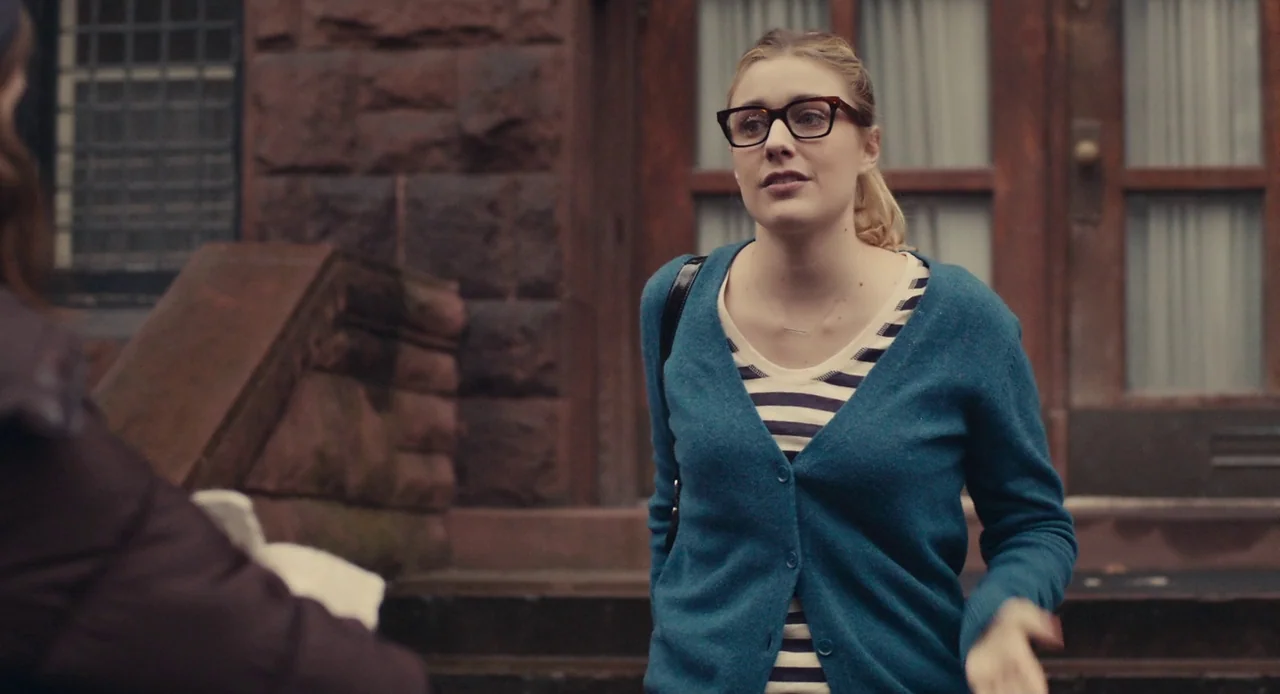 Mistress America (2015) movie still: medium — A stone brownstone building with a wooden entrance provides the backdrop. A woman standing on the…