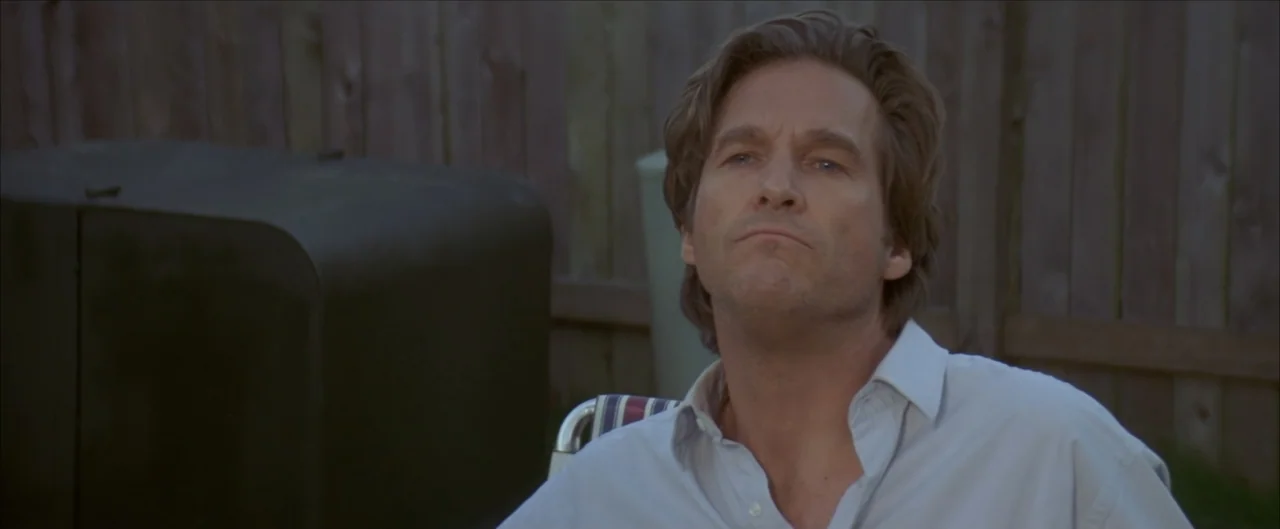 Arlington Road (1999) movie still: medium — A residential backyard features a wooden fence and a large dark object nearby. A man in a light shirt…