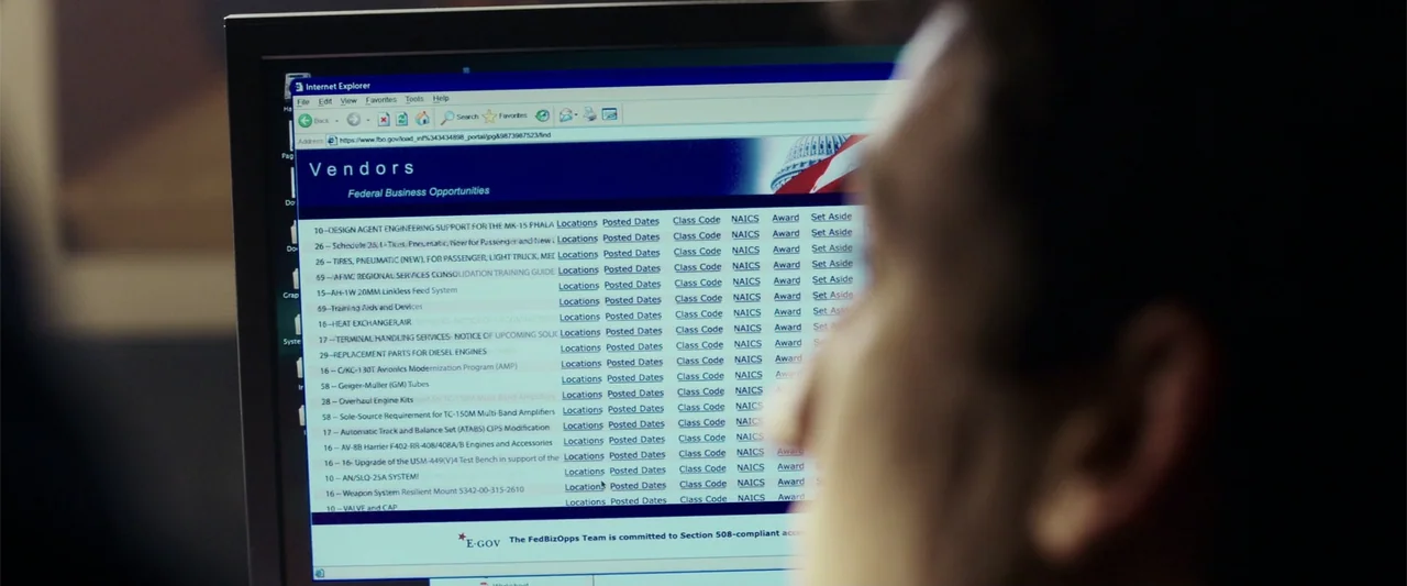 War Dogs (2016) movie still: medium — A dimly lit room contains a computer screen displaying a government vendor database website. A man is seated…