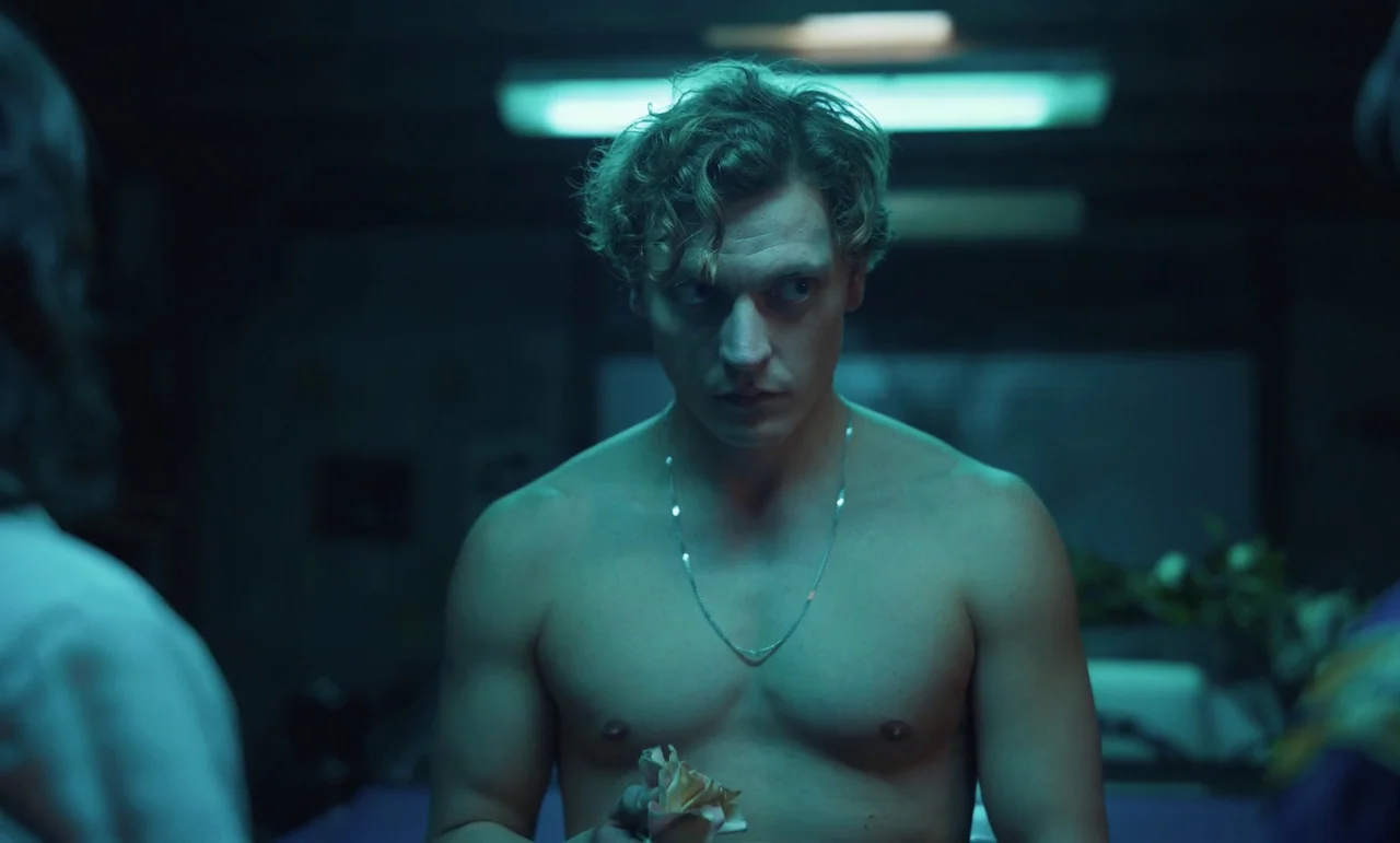 The Silent Twins (2022) movie still: medium — A dimly lit room is illuminated by a single cool-toned overhead fluorescent light. A shirtless young…