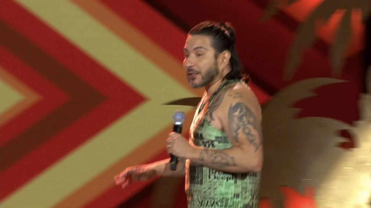 Felipe Avello: Doglover (2024) movie still: medium — A man stands before a large, patterned backdrop consisting of diagonal red and yellow stripes.…