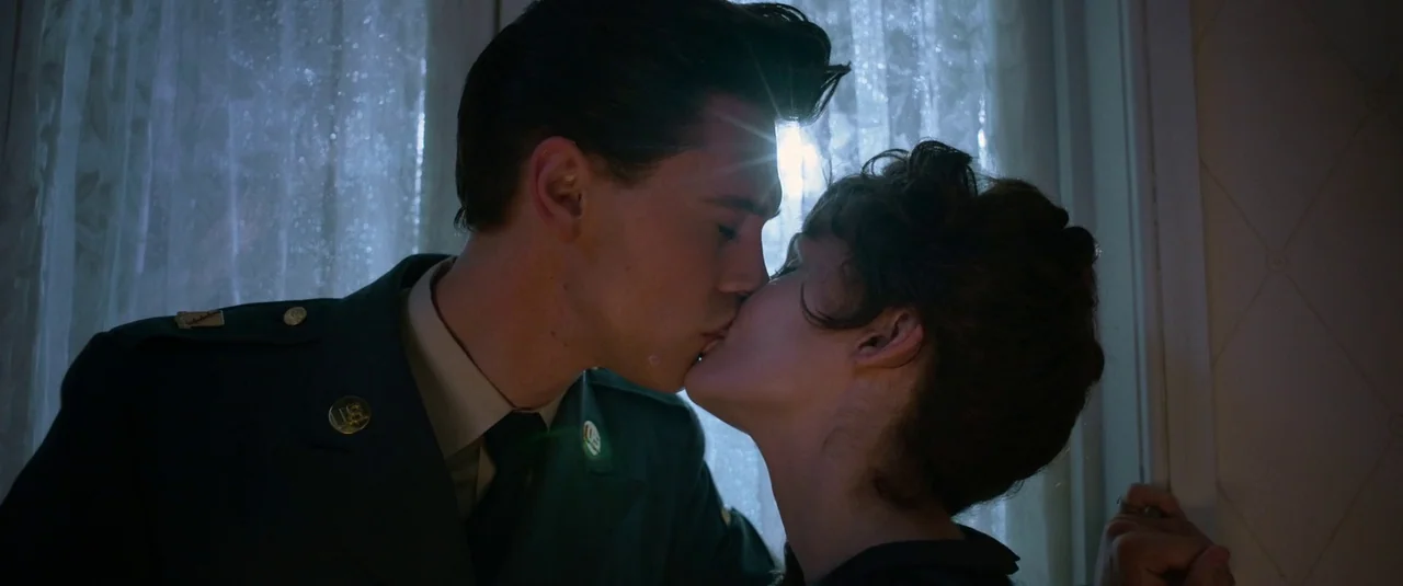 Elvis (2022) movie still: medium — A man and a woman kiss in front of a window with thin curtains. Sunlight flares behind the man's head while the…