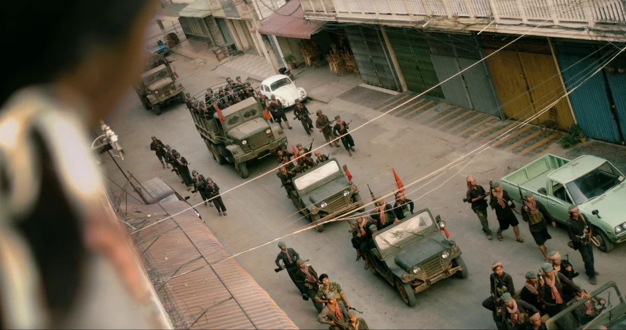 First They Killed My Father (2017) movie still: wide — A narrow city street is occupied by multiple olive drab military trucks and jeeps. A large…
