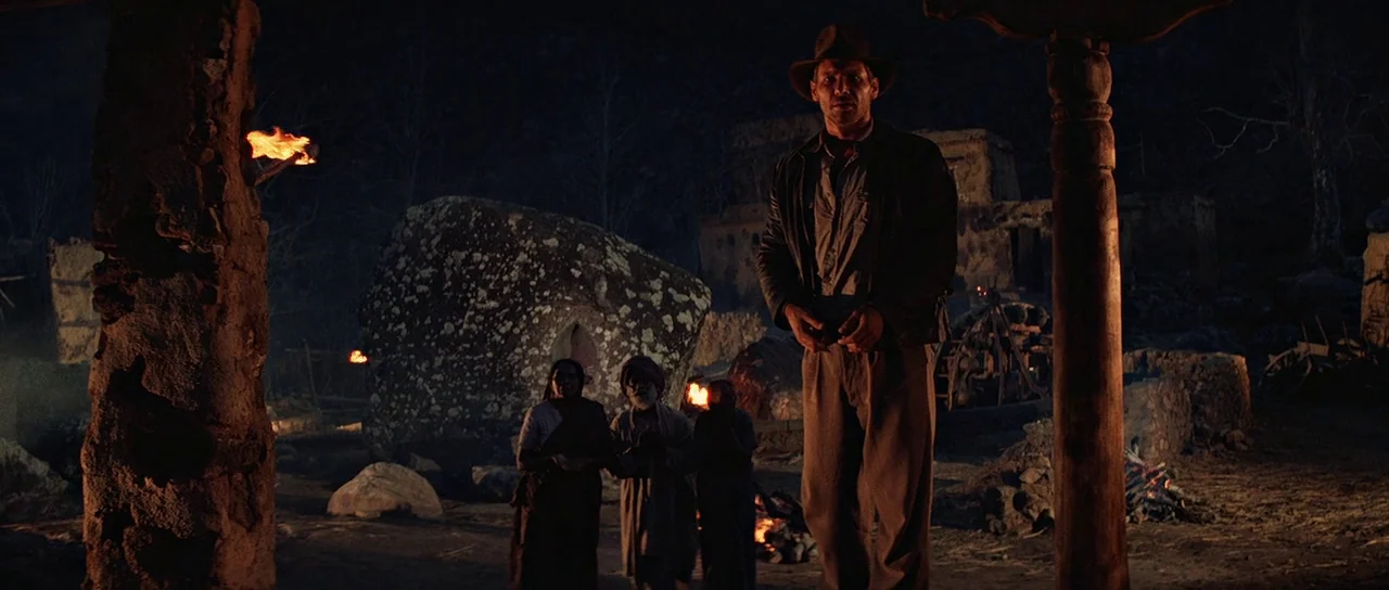 Indiana Jones and the Temple of Doom (1984) movie still: fullbody — A rural village at night is dimly lit by scattered firelight. A man in a hat…