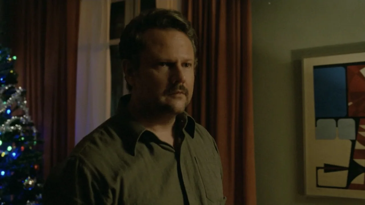 I'm Still Here (2024) movie still: medium — A domestic interior features dark curtains and an abstract framed print on the wall. A man in a green…