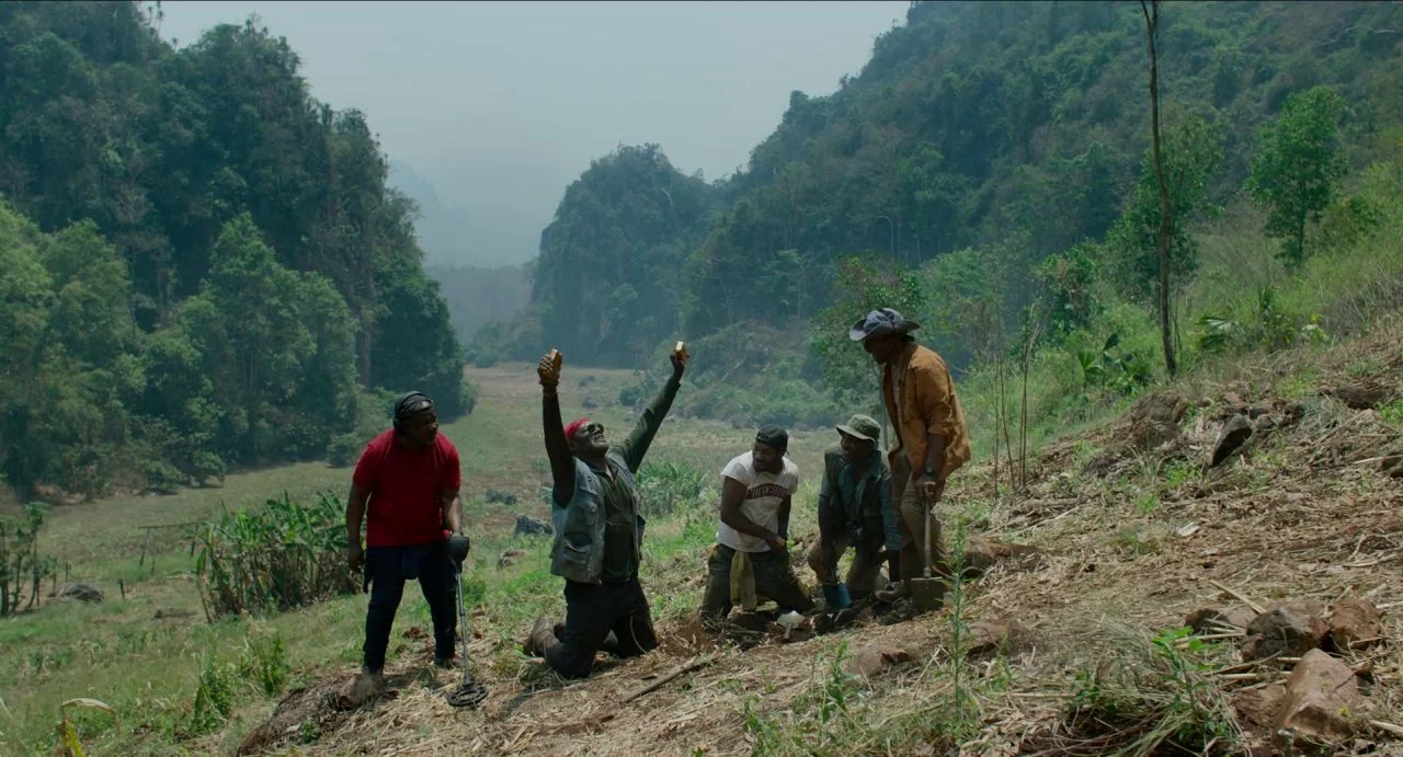 Da 5 Bloods (2020) movie still: wide — A group of five men stand on a hillside covered in dry grass and dirt. The center man kneels while holding…