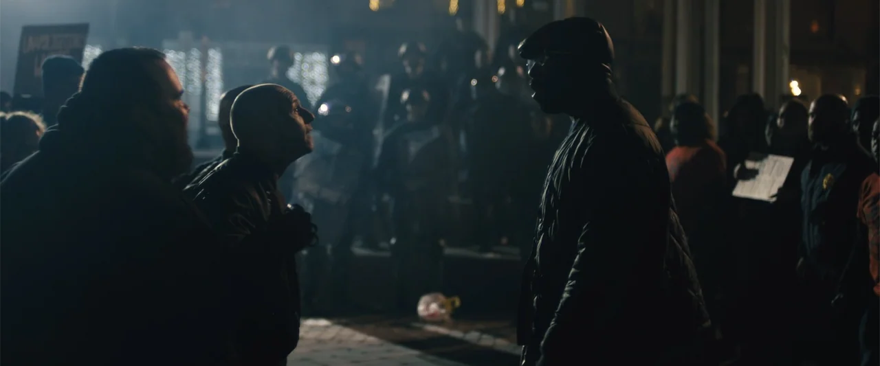 Skin (2009) movie still: medium — A nighttime protest features a crowd of people in front of a building entrance. Two men stand in the foreground…