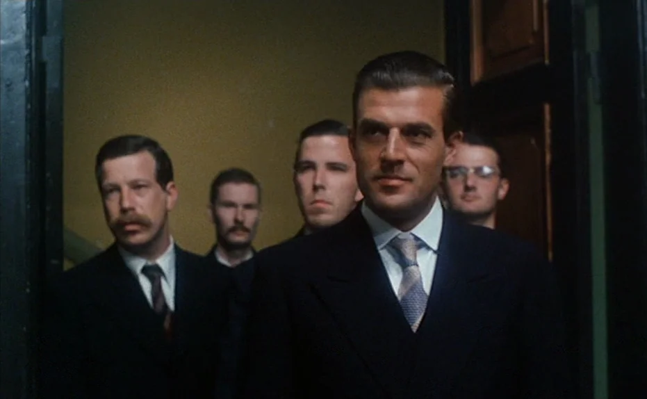 Soldier of Orange (1977) movie still: medium — A group of men in suits emerge from a dark doorway into a hallway. The men move forward toward the…