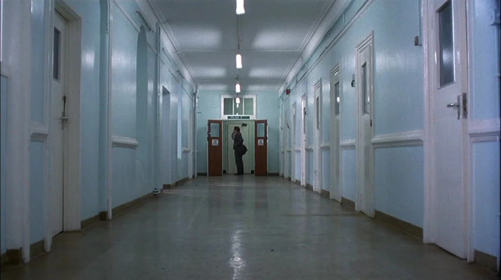 Velvet Goldmine (1998) movie still: wide — A long, sterile hospital hallway with pale blue walls and several identical wooden doors. A man in a dark…