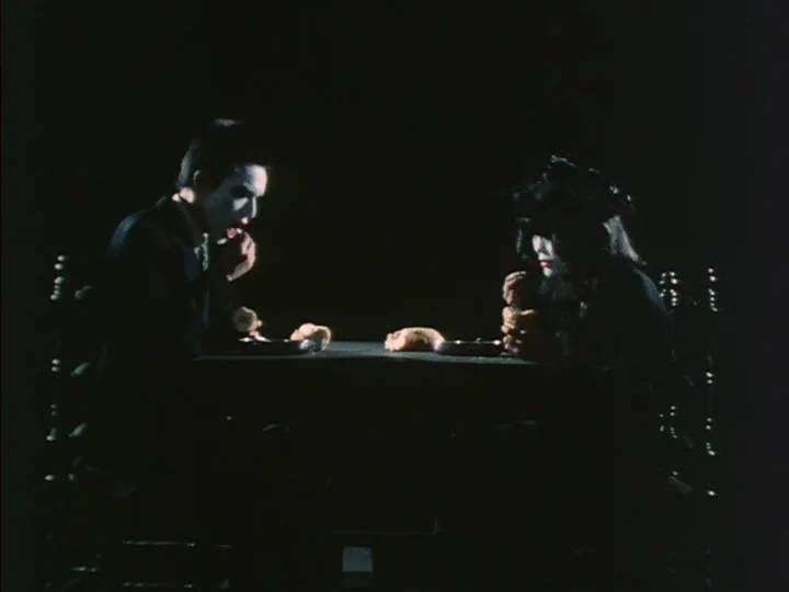 Trial (1975) movie still: medium — A dark, empty space features two figures sitting across from each other at a table. They are consuming bread in a…