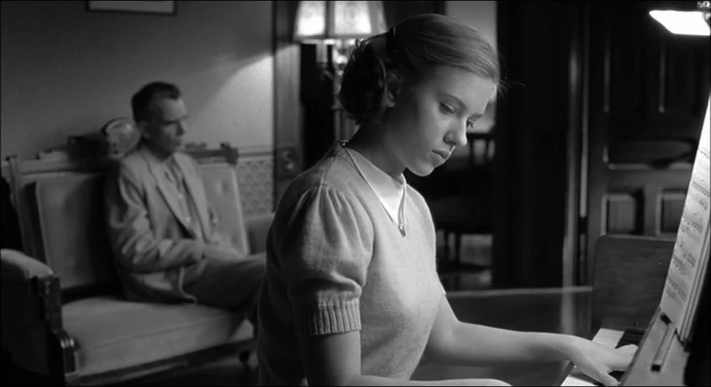 The Man Who Wasn’t There (2001) movie still: medium — A dimly lit room features a woman playing a piano in the foreground. A man sits on an…