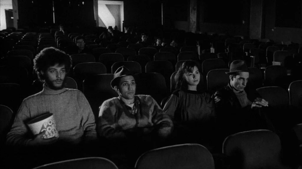 Stranger Than Paradise (1984) movie still: medium — Four people sit in a row of dark theater seats facing the screen. They watch the film as dim…
