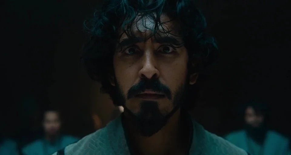 The Green Knight (2021) movie still: closeup — A dimly lit environment with a central male figure facing forward in darkness. Blurred figures are…