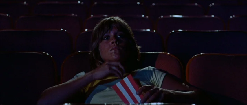 Messiah of Evil (1973) movie still: medium — A dark movie theater contains rows of empty cushioned seats in soft shadows. A young woman sits in the…