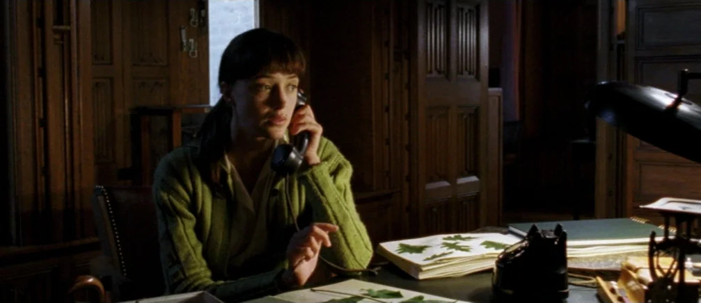 Innocence (2005) movie still: medium — A dark, wood-paneled room contains a desk cluttered with botanical samples and papers. A young woman in a…
