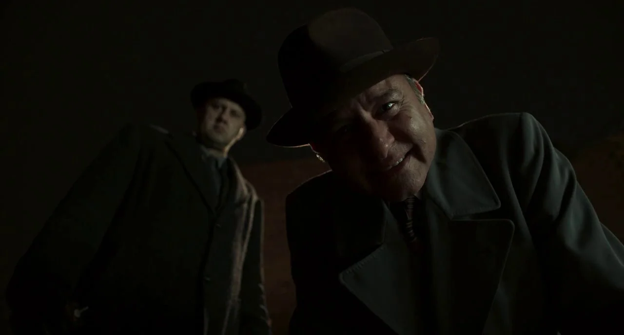 Motherless Brooklyn (2019) movie still: medium — Two men in 1950s attire stand in a dark, poorly lit room. They look directly down at the camera…