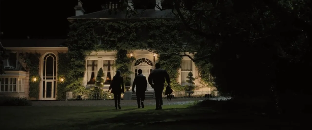 Abigail (2024) movie still: wide — A large two-story house with climbing vines at night sits behind a dark lawn. Three figures walk away from the…