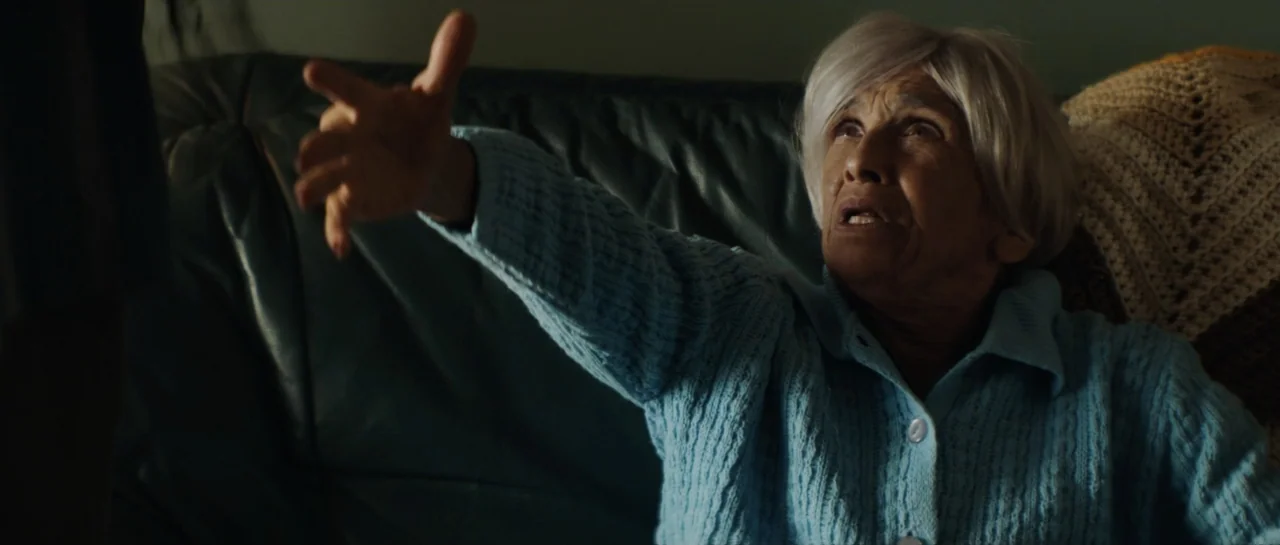 The Front Room (2024) movie still: medium — An elderly woman sits on a dark leather couch in a dimly lit room. She reaches out her right hand toward…